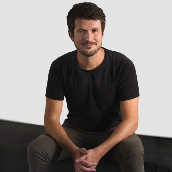Rodrigo Studart sitting with hands clasped, wearing a black T-shirt and dark pants against a simple light background.