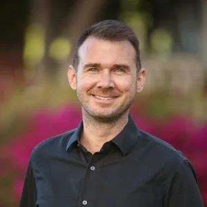 Sean Howell smiling outdoors while wearing a black shirt with blurred flowers behind him.