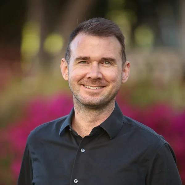 Sean Howell smiling outdoors while wearing a black shirt with blurred flowers behind him.