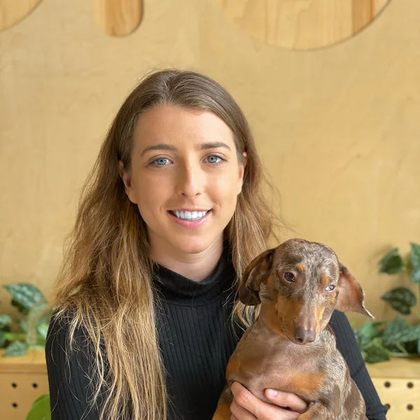 Shaina Denny with long hair wearing a black top, holding a small brown dachshund in front of a light wooden background.