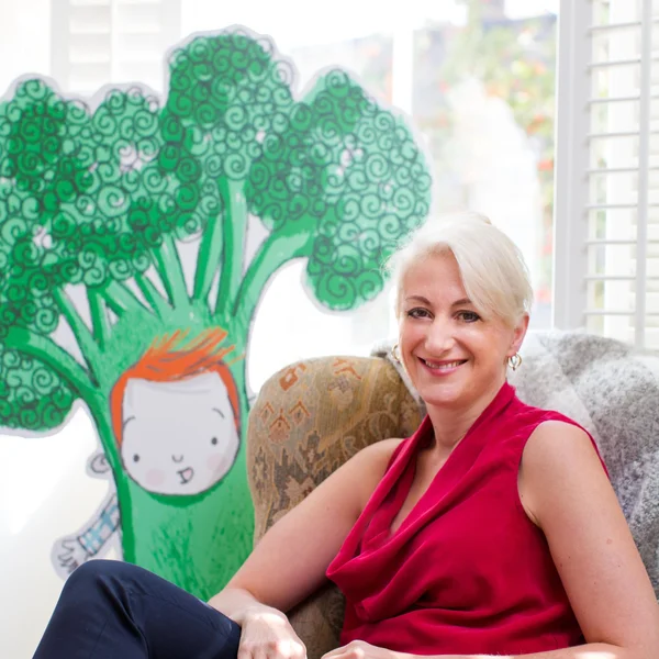 Sheena Macrae smiling in a chair beside a large illustrated tree character with a child.