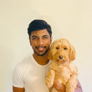 Shiv Sivakumar in a white shirt smiling while holding a small fluffy light-brown dog.