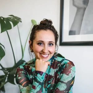 Smiling Susannah Schoolman in a colorful top resting her chin on her hand indoors.
