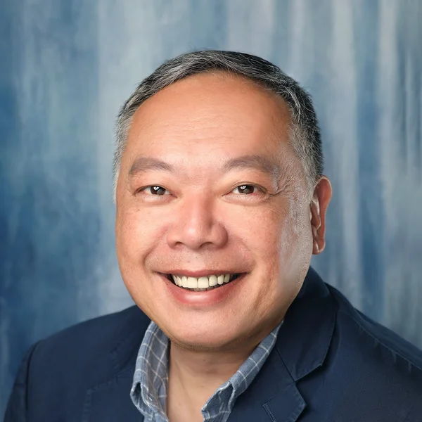 Smiling Thomas Tsang wearing a navy blazer and checkered shirt, posed in front of a blue textured background.