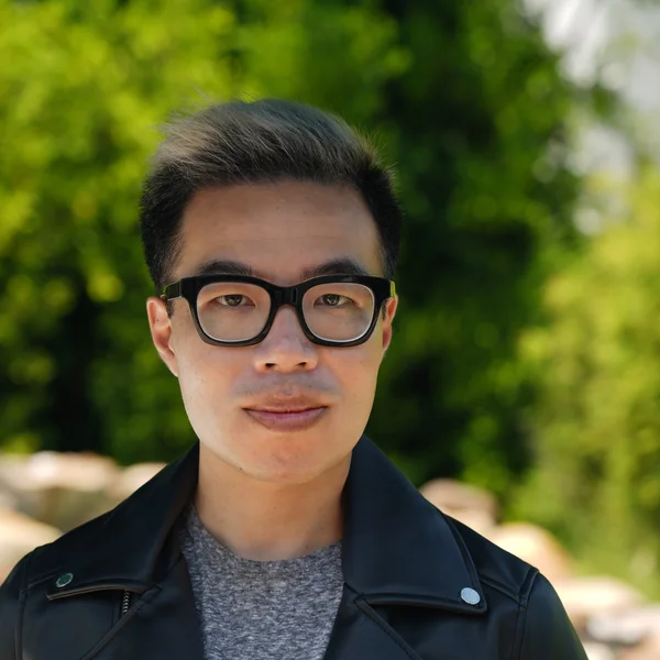 Tim Chan wearing black glasses and a black leather jacket, standing outdoors with greenery in the background.