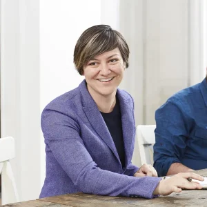 Smiling Vanessa Archambault in a purple blazer sitting at a table in a bright room.