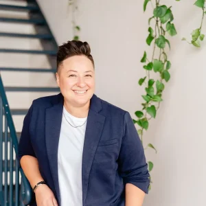 Smiling Vicky Pasche in a navy blazer and light shirt standing by a staircase with trailing green plants on the wall