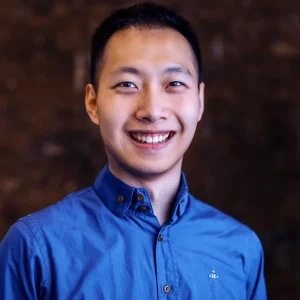 Smiling Yudi Ding wearing a blue button-up shirt against a dark blurred background.